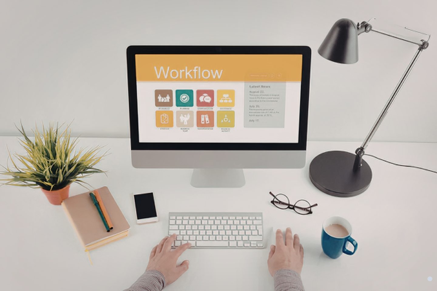 Person working at a desk with a computer displaying a workflow dashboard, surrounded by office items like a notebook, phone, and coffee.